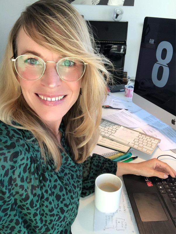 Eine Frau mit Brille sitzt l&auml;chelnd an einem Schreibtisch und tippt auf einer Tastatur, w&auml;hrend eine Tasse Tee vor ihr steht.