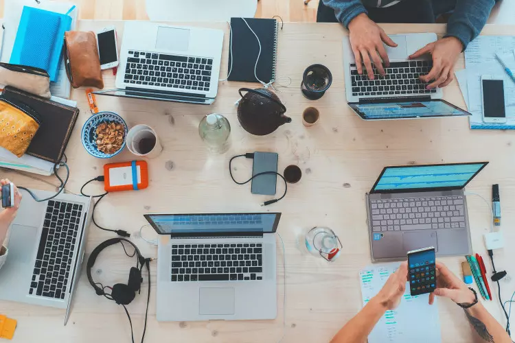 Ein Arbeitsplatz mit mehreren Laptops, pers&ouml;nlichen Gegenst&auml;nden und Personen, die an verschiedenen Aufgaben arbeiten.